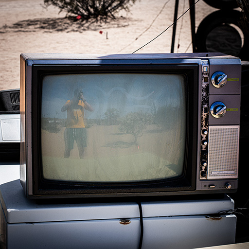 A photo of an old CRT television sitting outside in the desert at the Noah Purifoy Outdoor Desert Art Museum in Joshua Tree, California. My reflection is visible in the left third of the monitor, holding the camera to take the photo.